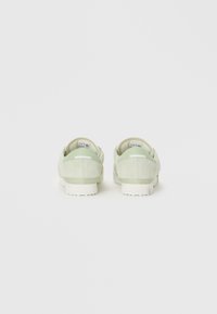 Un par de zapatillas de ante verde claro mostradas desde atrás sobre un fondo blanco, destacando el diseño del talón y la suela.
