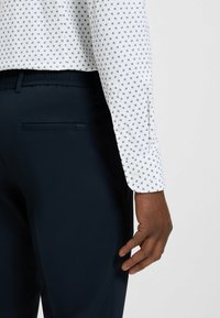 Witte button-up shirt met kleine zwarte patronen, gecombineerd met marineblauwe broek met een zijzak met rits en een elastische tailleband.