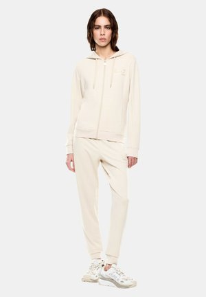 Donna che indossa una giacca con cappuccio beige con zip, pantaloni jogger abbinati e sneakers bianche, in piedi contro uno sfondo semplice.