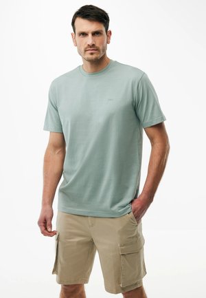 Ein Mann trägt ein hellgrünes kurzärmeliges T-Shirt und beige Cargo-Shorts und steht mit einer Hand in der Tasche vor einem weißen Hintergrund.