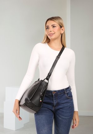 Sac à épaule en cuir noir au design souple, avec une poche zippée et une sangle réglable. Porté sur un haut blanc à manches longues et un jean bleu.