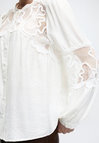 Hvit bluse med blonderdetaljer på skuldre og ermer, med knapper trukket i stoff og puffy lange ermer.