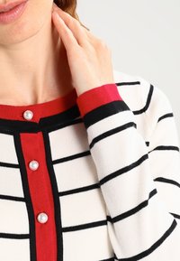 Randig kofta i vitt, svart och rött. Har rund halsringning, pärlliknande knappar och ribbade ärmslut. Mjuk textur med en figurvänlig design.