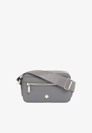 Sac bandoulière rectangulaire gris avec poche zippée à l'avant, tirette de zip argentée et large bandoulière réglable embossée du nom de la marque "LACOSTE".