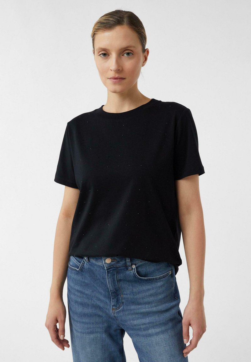 Woman with pulled-back hair wearing a black short-sleeve t-shirt and blue jeans, standing against a plain white background.