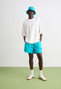 Camiseta blanca de gran tamaño, pantalones cortos turquesa y sombrero de cubo a juego. Zapatillas ligeras con acentos verdes y calcetines blancos. Look sencillo y deportivo.