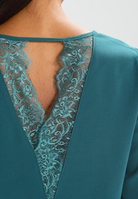 Schiena della scollatura di un indumento verde smeraldo con bordo in pizzo floreale che lascia intravedere la pelle della parte superiore della schiena e una sottile tracolla sopra.