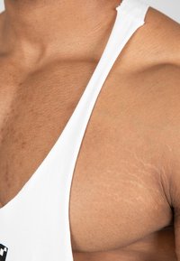 Witte tanktop met brede bandjes, die de gestructureerde huid en schoudercontouren toont, met de nadruk op de halslijn en het borstgebied.