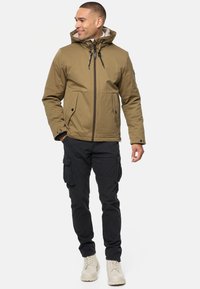 Giacca a cappuccio marrone con zip frontale, due ampie tasche laterali e pantaloni cargo neri. Il modello indossa stivali beige; tessuto morbido e vestibilità strutturata.