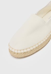 Balti drobės espadrilės su suapvalintu pirštu, jūtinio vijoklio pado, ir matomomis siūlėmis palei kraštą. Lygus paviršius ir paprastas dizainas.