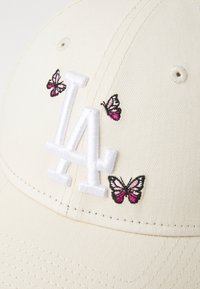 Kremfarget baseballcaps med en hvit "LA"-logo og tre broderte sommerfugler i rosa, svart og hvitt. Teksturert stoff.