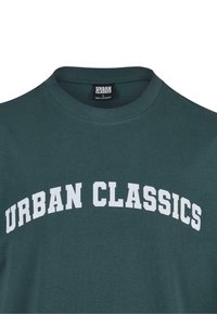 Sötétzöld pamut póló, felsőkerek nyakkivágással és az "URBAN CLASSICS" felirattal, vastag fehér betűkkel a mellkason.