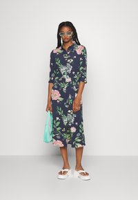 Vestido floral azul-marinho com mangas até ao cotovelo, botões e cintura com laço; combinado com sandálias brancas de plataforma e uma bolsa azul-claro.