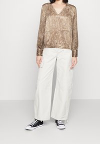 Blouse marron à manches longues et décolleté en v avec des rayures verticales, associée à un pantalon cargo beige ample et des baskets basses noires.