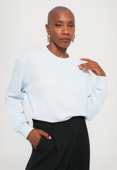 Lyseblå genser med lange ermer, med en sirkulær grafikk i gulltekst. Stilt med svarte høyhalsbukser.