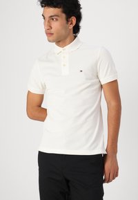 Polo shirt blanc à manches courtes avec une patte de boutonnage à trois boutons et un petit logo sur la poitrine. Le tissu a une finition lisse et légèrement texturée.