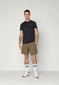 Μαύρο αθλητικό μπλουζάκι και ελιόχρωμο σορτς. Το μοντέλο φοράει λευκά αθλητικά παπούτσια με ανοιχτό καφέ σόλες και ριγέ λευκές και μαύρες κάλτσες.