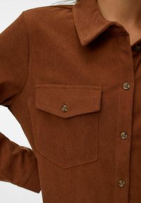Chemise en velours côtelé marron avec col, devant boutonné et poche poitrine. Le tissu présente une texture à côtes verticales et des boutons marron.