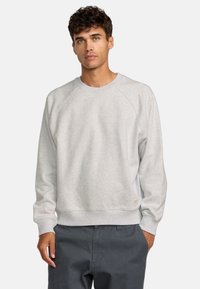 Sweatshirt gris clair avec des manches raglan, tissu texturé, col et poignets côtelés, et un petit logo en patch sur l'ourlet. Porté par-dessus un pantalon sombre.