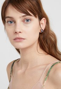 Orecchini quadrati oro con accenti di glitter colorati. La modella ha i capelli castano chiaro e indossa un top leggero e trasparente con spalline verdi.