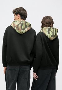 Fekete kapucnis pulóverek terepmintás kapucnival, laza szabásúak, bordázott mandzsettával, sötétszürke farmerrel viselve, hátulnézetből.
