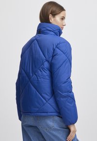 Blå puffjacka med hög krage, med en quiltad design och ribbade ärmar. Slät, glänsande textur med en figurnära siluett.