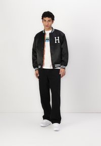 Jachetă de vară din satin negru cu dungi albe și litera "H," peste un tricou grafic alb. Asortată cu pantaloni negri și adidași albi.