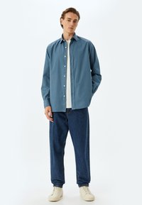 Camisa de botones azul claro con cuello; se lleva sobre una camiseta blanca y se combina con jeans de denim oscuro y zapatillas crema.
