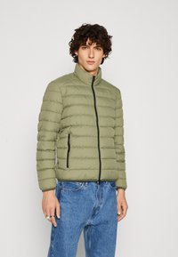 Giacca imbottita verde oliva con colletto alto, zip frontale e cuciture orizzontali. Presenta due tasche laterali con zip nere.