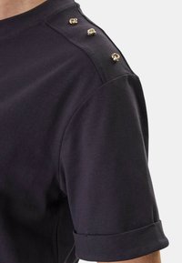 Gros plan sur une chemise noire à manches courtes avec trois boutons décoratifs torsadés en or sur la couture de l'épaule.