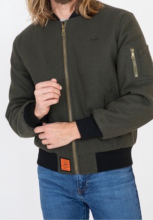 Mann trägt dunkelgrüne Bomberjacke mit Reißverschluss, schwarzen Bündchen und blauer Jeans, zieht Ärmel mit sichtbaren Händen zurecht.