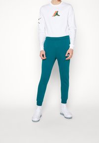 Vit långärmad skjorta med en färgglad logotyp på bröstet, kombinerad med turkosa joggingsbyxor och vita sportskor.