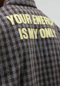 Bruin geruit overhemd met een gestructureerde stof en een gedurfde gele tekst op de achterkant: "YOUR ENERGY IS MY ONLY."