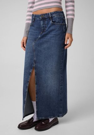 Mannequin portant une longue jupe en jean bleue fendue à l’avant, un pull court à rayures, des chaussettes blanches et des mocassins foncés.