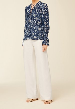 Woman wearing a navy floral long-sleeve blouse with lace-up neckline paired with high-waisted white wide-leg trousers and beige heels.