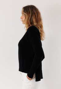 Maglione nero lavorato a maglia con maniche lunghe, bordo a coste e fessure laterali, indossato sopra pantaloni bianchi. Texture liscia. Modella vista di lato.