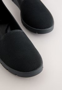 Chaussures slip-on noires avec dessus en tissu mesh respirant et semelles épaisses en caoutchouc, exposées sur une surface claire et unie.
