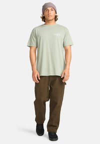 Lysegrønn bomull t-skjorte med korte ermer og et brystlogo, kombinert med brune cargo-bukser og svarte joggesko. Myk tekstur.