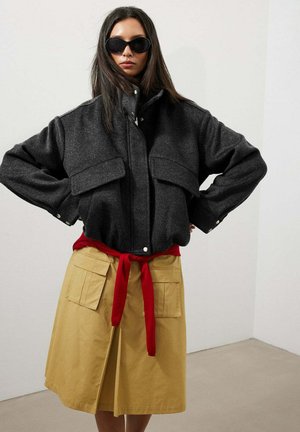 Mujer con cabello largo y oscuro, que lleva gafas de sol negras, una chaqueta holgada gris oscuro, un cinturón rojo y una falda beige con bolsillos delanteros.