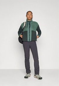 Kurtka polarowa z zielonym wzorem blokowym, zapinana na zamek z przodu, z regulowanym kapturem, zestawiona z ciemnymi spodniami turystycznymi i szarą obuwiem trekkingowym.