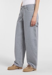 Personne portant un pantalon ample rayé verticalement gris et blanc avec une poche latérale, associé à des chaussures beiges à lacets et un haut blanc.