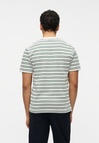 Άνδρας με σγουρά μαλλιά φοράει λευκό κοντομάνικο πουκάμισο με λεπτές μαύρες και πράσινες οριζόντιες ρίγες, στέκεται γυρισμένος προς τα πίσω σε λευκό φόντο.