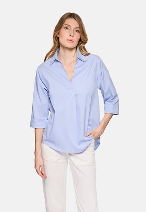 Mujer de cabello castaño claro que lleva una blusa azul claro de manga tres cuartos y pantalones blancos, de pie con una mano en el bolsillo.