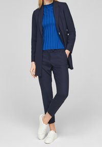 Marinblå blazer över en blå ribbad tröja, kombinerad med marinblå avsmalnande byxor och vita sneakers. Slätt tyg med rena linjer och skräddarsydd passform.