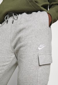 Personne portant un pantalon de survêtement gris Nike avec une poche latérale et un sweat à capuche vert, une main dans la poche, montrant les cordons de serrage et le détail du logo.