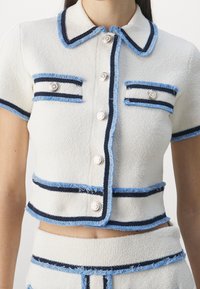 Bílý pletený top s krátkým rukávem, který má modré a námořnické pruhy, dekorativní knoflíky a třepené okraje kolem límce a kapes.