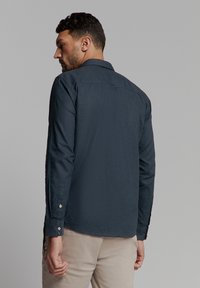 Marineblauw overhemd met lange mouwen, een knoop-neklijn, gestructureerde stof en knopen bij de polsen. Bevat een rugneklijn detail.