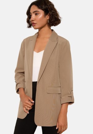 Frau mit lockigem dunklem Haar, die einen beigen Blazer über einem weißen Oberteil und schwarzen Hosen trägt und vor einem schlichten Hintergrund posiert.