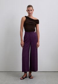 Brown off-shoulder top in soft fabric, paired with high-waisted, wide-leg purple pants featuring two gold buttons and side pockets.