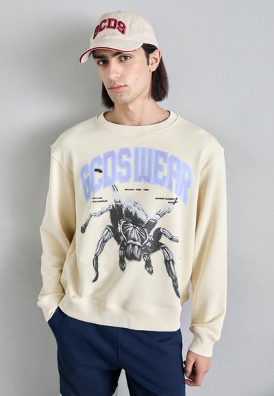 Sweatshirt beige avec un motif d'araignée, comportant un texte bleu et noir disant "GCDSWEAR." Le modèle porte un short bleu marine et une casquette beige avec des lettres rouges.